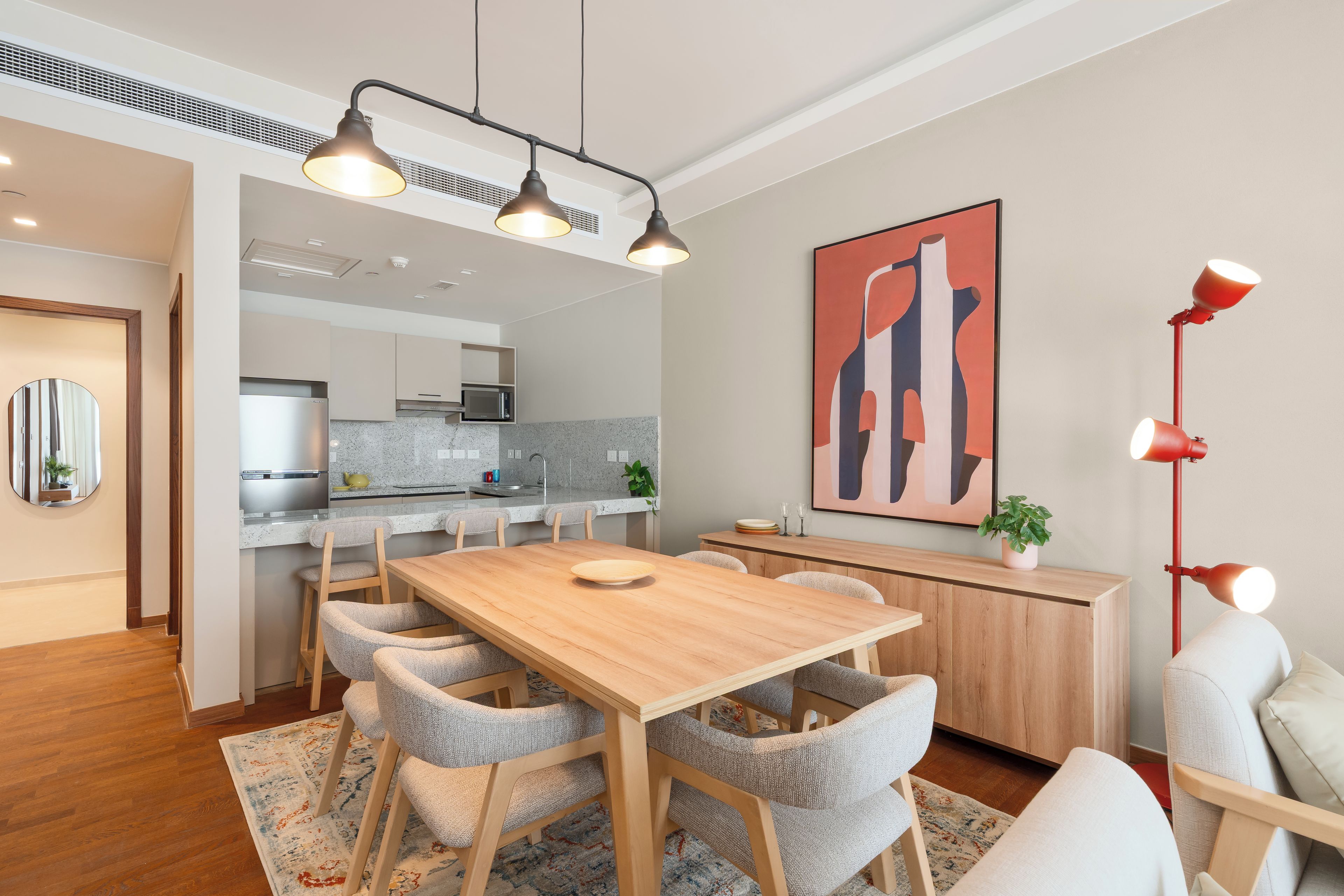 A modern dining area with a wooden table, grey upholstered chairs, a patterned rug, a red floor lamp, and a kitchen in the background.