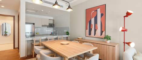 A modern dining area with a wooden table, grey upholstered chairs, a patterned rug, a red floor lamp, and a kitchen in the background.