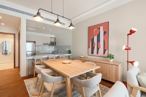 A modern dining area with a wooden table, grey upholstered chairs, a patterned rug, a red floor lamp, and a kitchen in the background. - Brassbell Sheikh Zayed Aeon Towers (6th of October City)