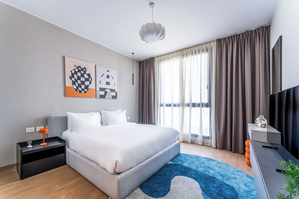 Una habitación de hotel moderna con una cama grande, una mesita de noche con lámpara, un televisor de pantalla plana y un ventanal con cortinas.