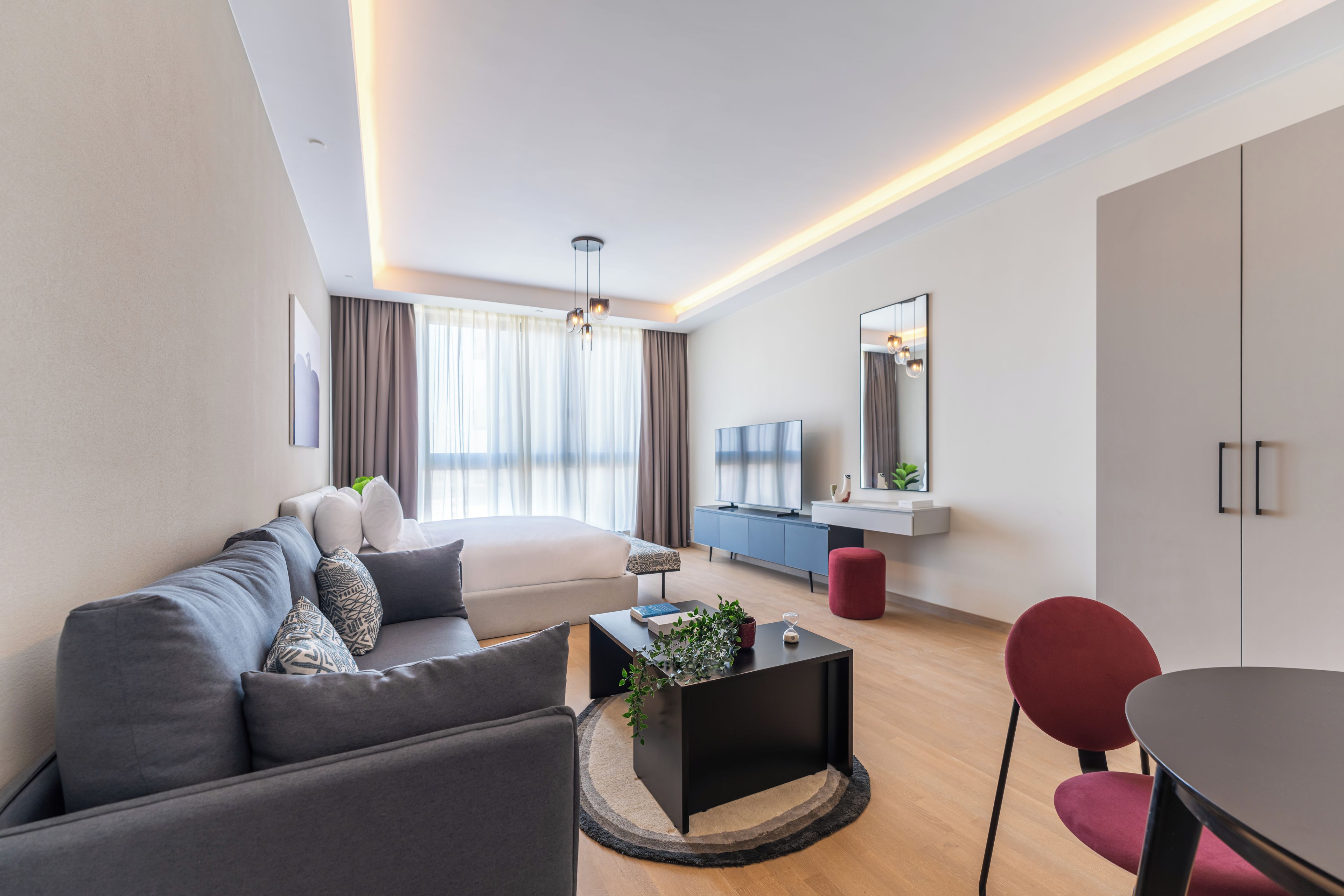 Sala de estar moderna com sofá, mesa de centro e cadeira vermelha. Há uma televisão, um espelho e uma janela com cortinas.