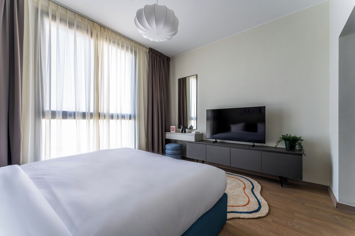 Una habitación de hotel moderna con una cama grande, un televisor de pantalla plana, un escritorio pequeño y un ventanal con cortinas.