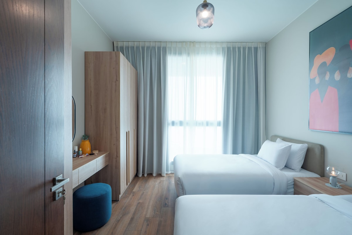 Una habitación de hotel moderna con dos camas, un armario de madera, un reposapiés azul y un ventanal con cortinas.