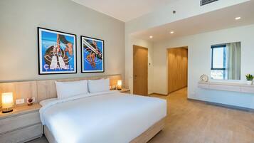 A modern hotel room with a large bed, two wall-mounted artworks, a bedside table with a lamp, and a window with a view.