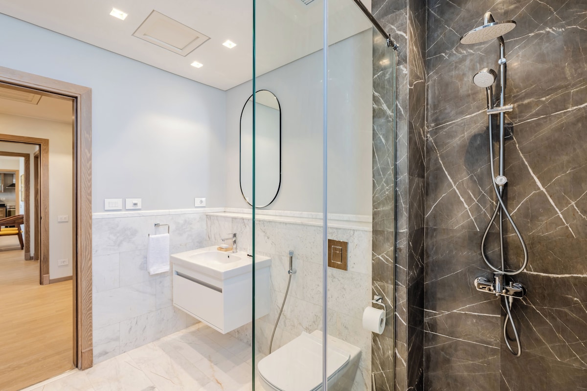 A modern bathroom with a glass-enclosed shower, a white sink, and a marble-tiled wall.
