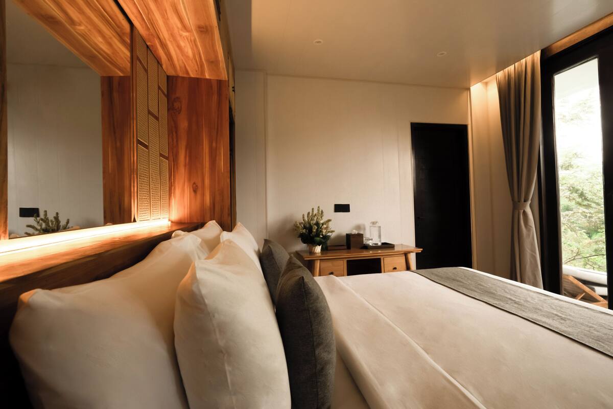A modern bedroom with a large bed, wooden headboard, bedside table, and a view of greenery outside.