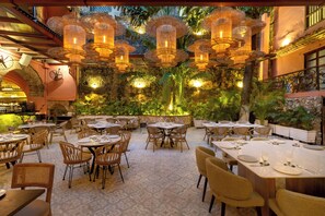 Un espace de restauration en plein air avec des tables rondes et des chaises en osier, entouré de verdure et agrémenté de guirlandes lumineuses décoratives.