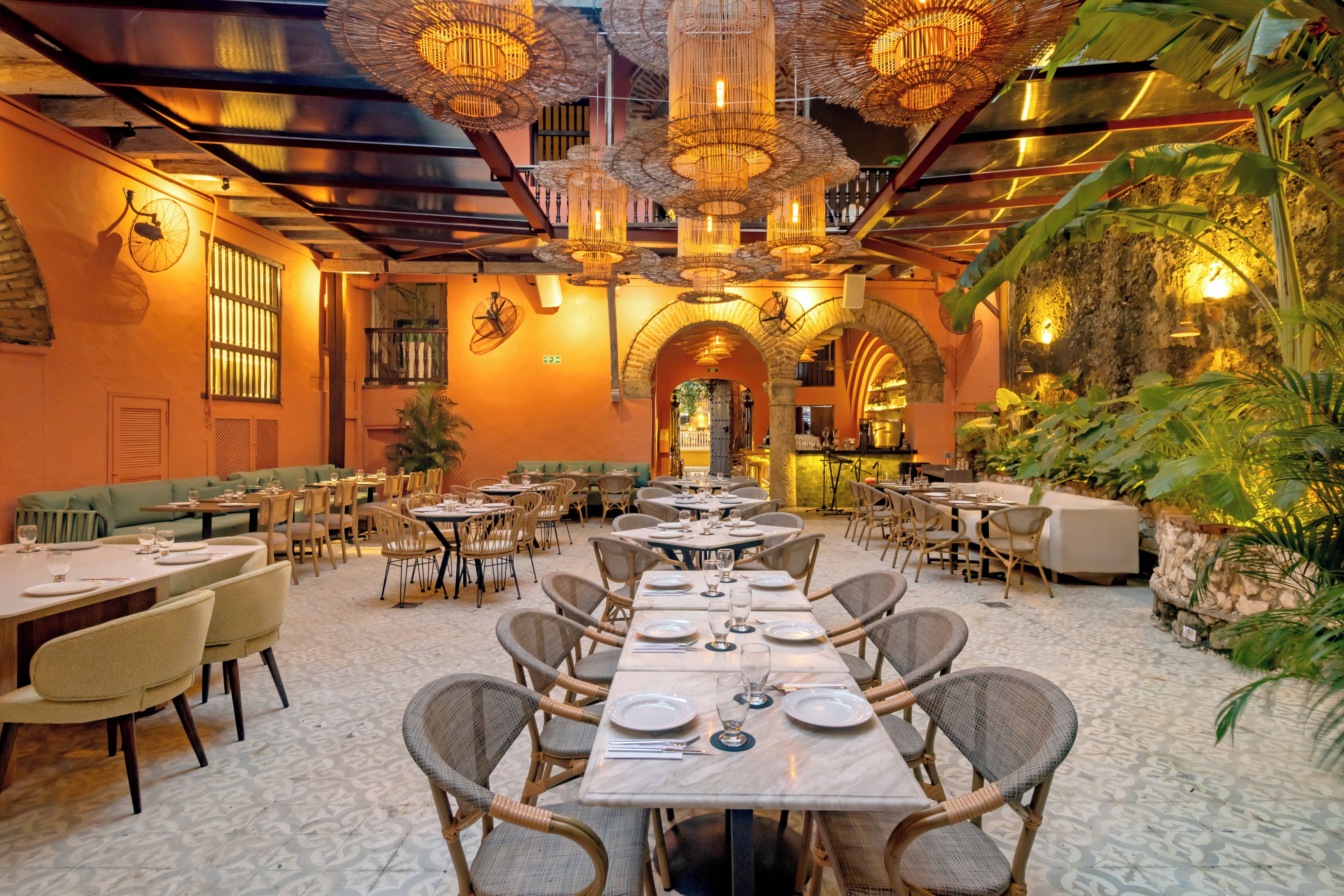 A restaurant with multiple dining tables and chairs, featuring a tropical theme with plants and wicker furniture.