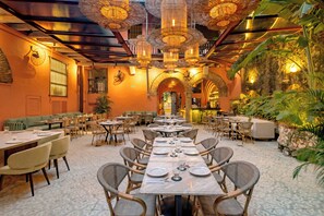Een restaurant met meerdere eettafels en stoelen, met een tropisch thema, planten en rieten meubels.