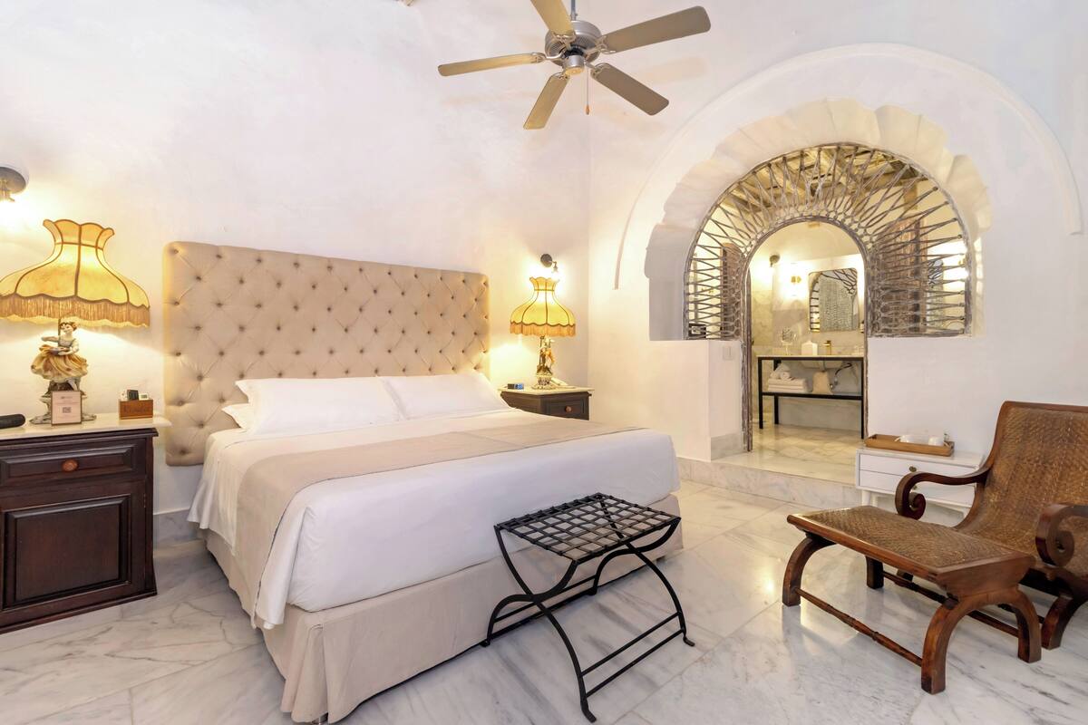 Une chambre à coucher avec une tête de lit capitonnée, un lit recouvert de draps blancs, une table de chevet avec une lampe, une petite table, une chaise et une porte en arc donnant sur une salle de bain.