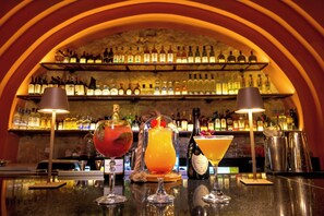 Um balcão de bar com três bebidas, uma estante de garrafas ao fundo e uma parede de pedra.
