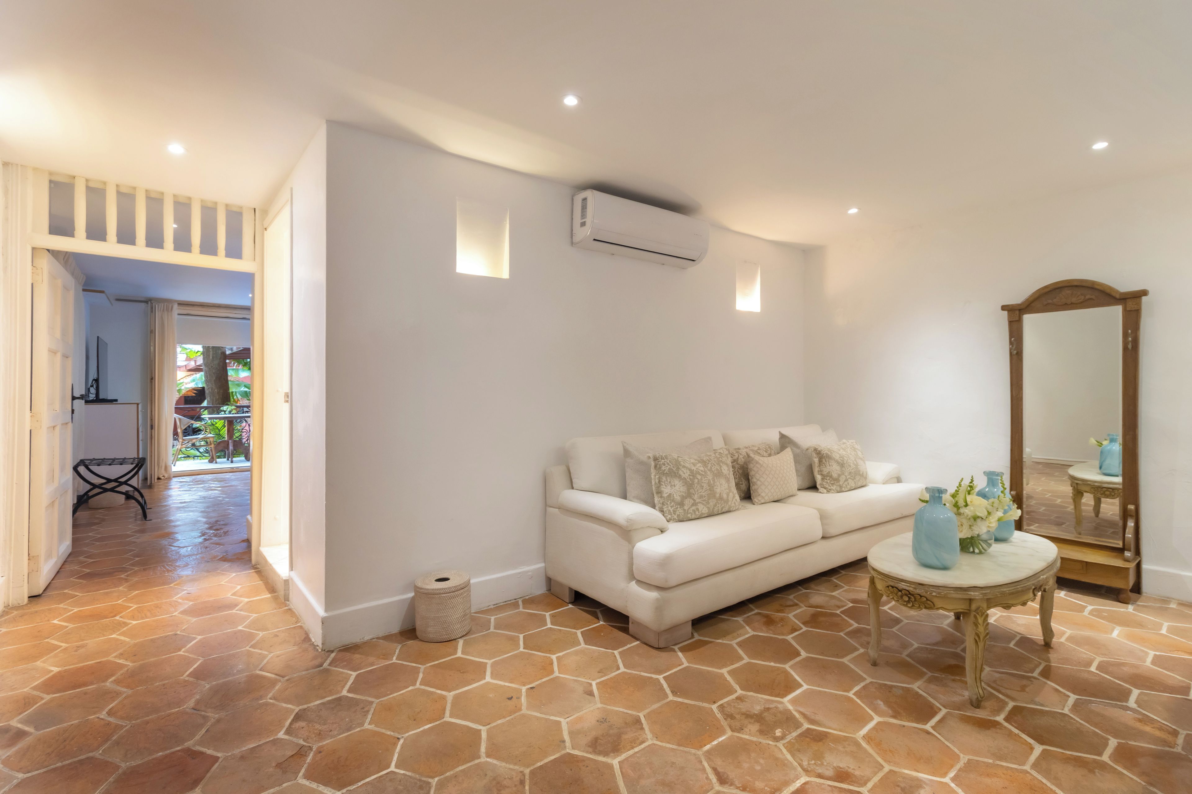 A modern living room with a white sofa, a round table with a vase, a large mirror, and a stone-tiled floor.