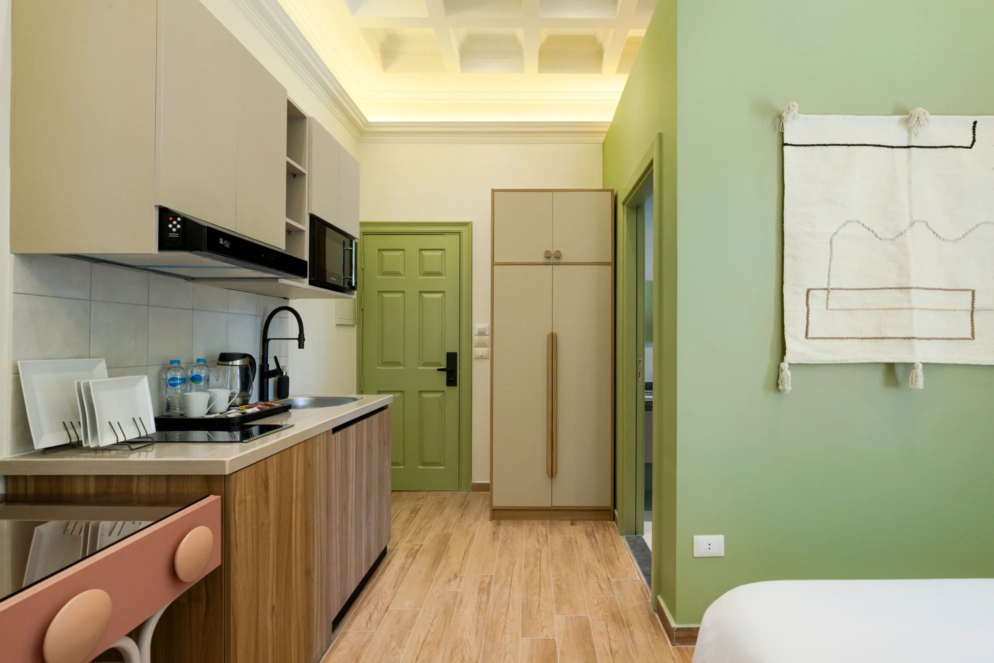 A compact kitchen with a microwave, sink, and cabinets, a green door, and a bed with a simple headboard.