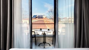 A room with a bed, a table, and a view of a building with solar panels.