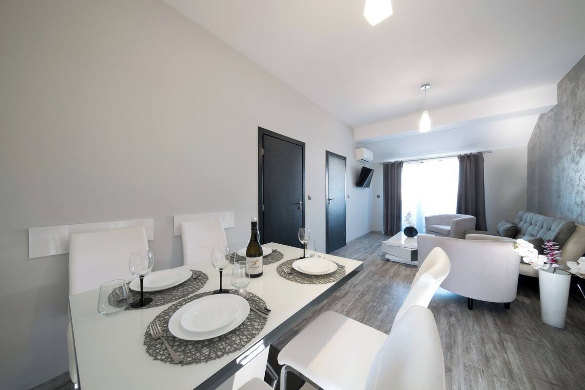 A modern dining area with a white table set for two, a bottle of wine, and a flat-screen TV mounted on the wall.