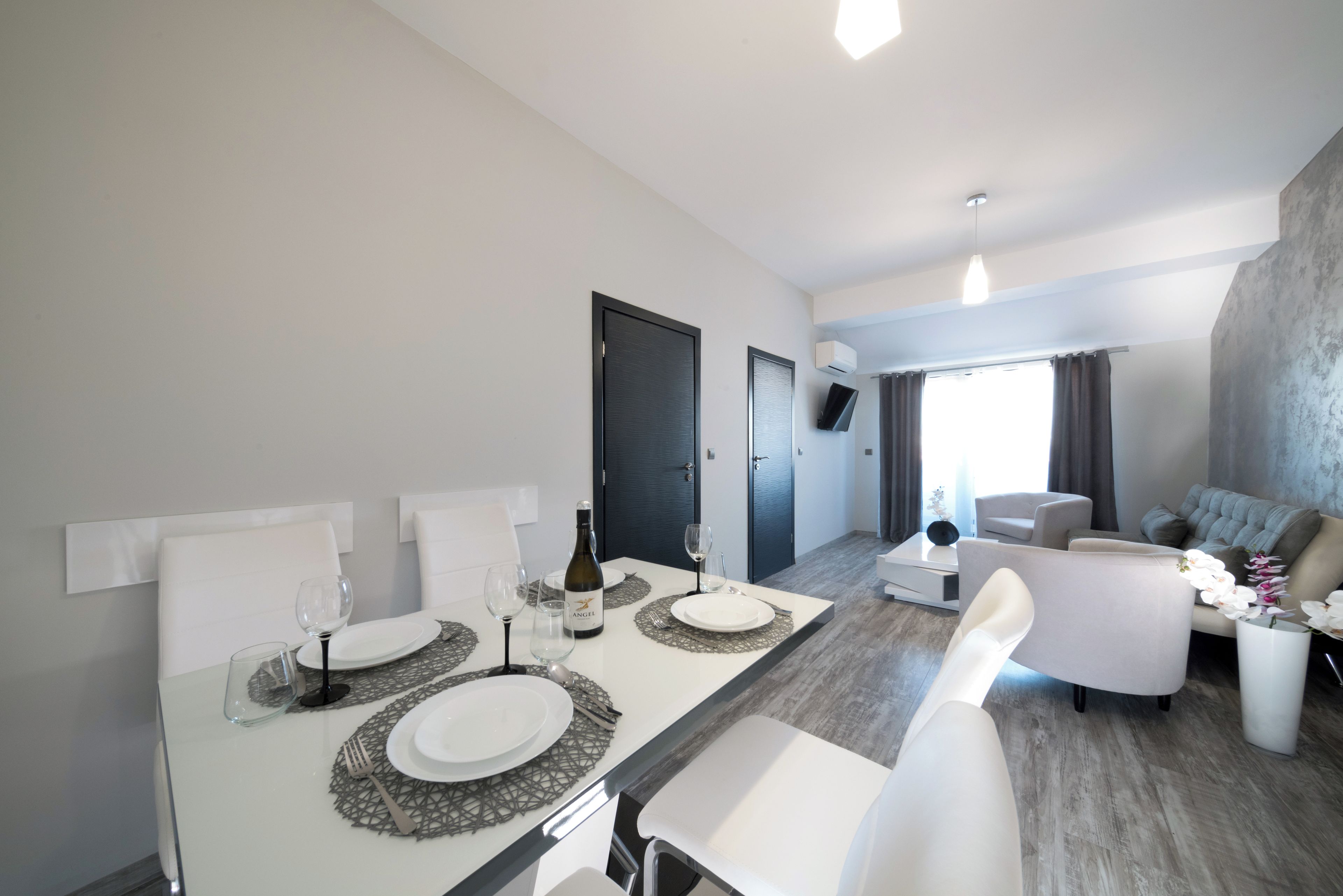 A modern dining area with a white table set for two, a bottle of wine, and a flat-screen TV mounted on the wall.