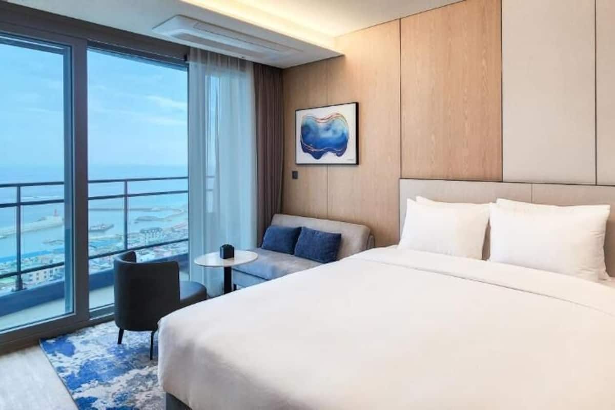 A hotel room with a large bed, a small table, a chair, and a view of the city.