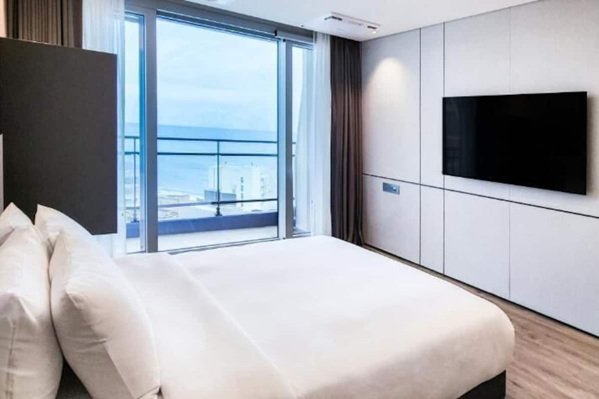 A hotel room with a bed, a TV mounted on the wall, and a large window offering a view of the city.