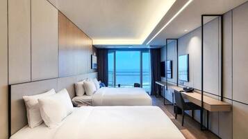 A modern hotel room with two beds, a desk, and a large window offering a view of the sea.