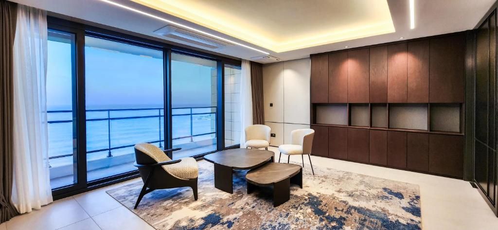 A modern hotel room with a large window offering a view of the sea, a seating area with a coffee table, and a dark wood panel wall.
