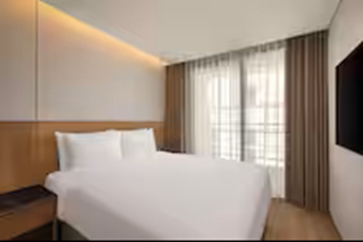 Habitación de hotel con una cama grande, una mesita de noche, un televisor de pantalla plana montado en la pared y una ventana con cortinas.