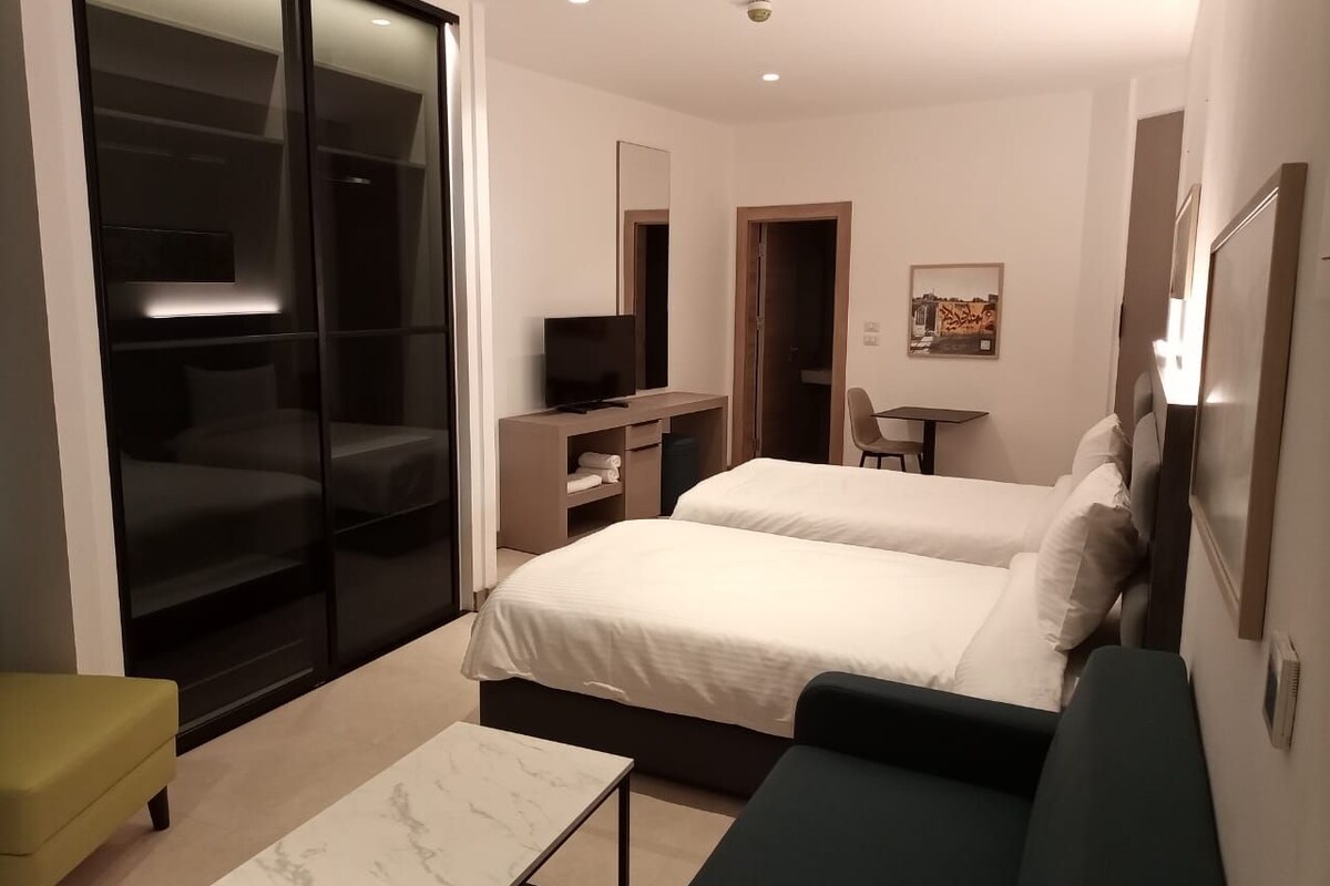 A modern hotel room with two beds, a desk, a chair, and a sliding glass door leading to a balcony.