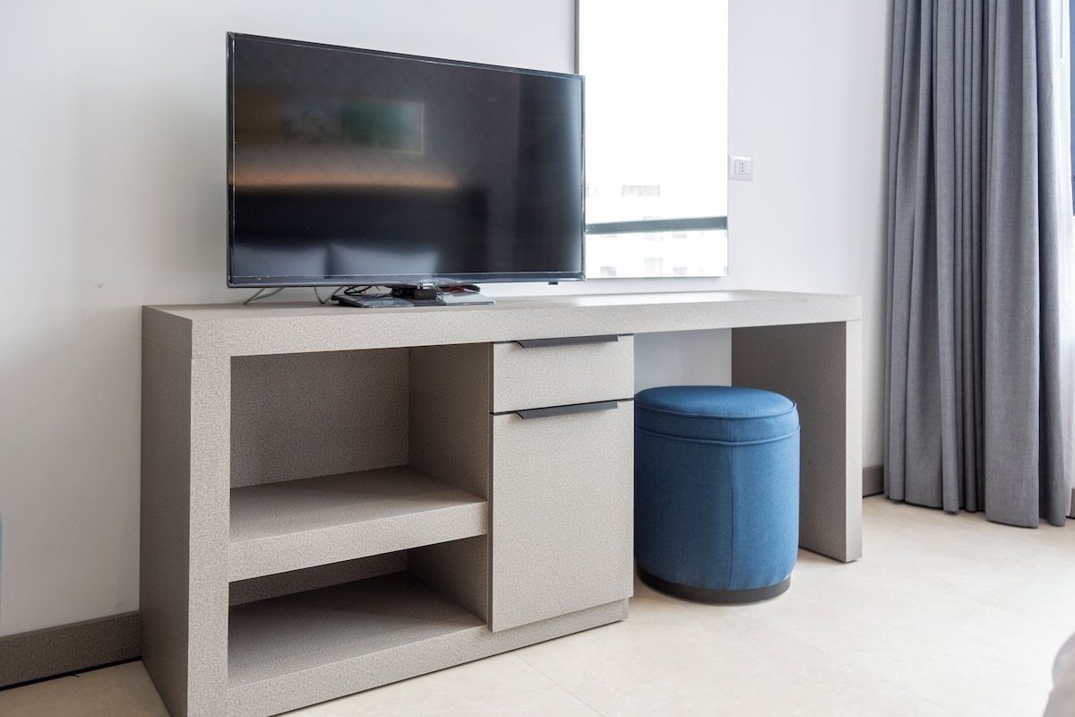 A modern, minimalist room with a flat-screen TV on a grey console, a small blue ottoman, and a window with grey curtains.