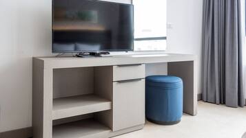 A modern, minimalist room with a flat-screen TV on a grey console, a small blue ottoman, and a window with grey curtains.