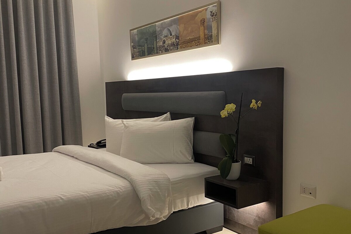 A modern bedroom with a bed, a bedside table, a nightstand, a potted plant, and a painting on the wall.