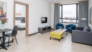 Une chambre d’hôtel moderne équipée d’un lit, d’une télévision, d’un coin repas avec une table et des chaises, d’un canapé et d’une grande fenêtre donnant sur la ville.