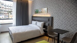Une chambre d’hôtel avec un lit, un bureau, deux chaises, un pouf vert et une fenêtre avec des rideaux.