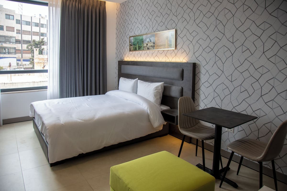 Une chambre d’hôtel avec un lit, un bureau, deux chaises, un pouf vert et une fenêtre avec des rideaux.