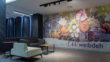 A modern lobby with a mural of flowers and the name 'Weibdeh' on the wall, a seating area with a sofa and tables, and a high ceiling with recessed lighting.