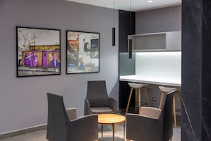 Un área de espera moderna con una mesa de madera y cuatro sillas, dos obras de arte enmarcadas en la pared y un mostrador blanco con una columna de mármol negro.