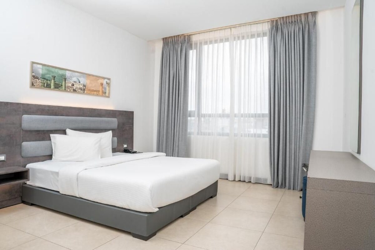 A modern hotel room with a large bed, a nightstand, a desk, and a window with curtains.
