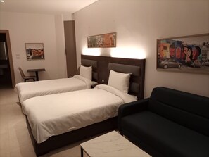 Une chambre d’hôtel avec deux lits, un canapé de couleur sombre, une petite table et des œuvres d’art accrochées aux murs.