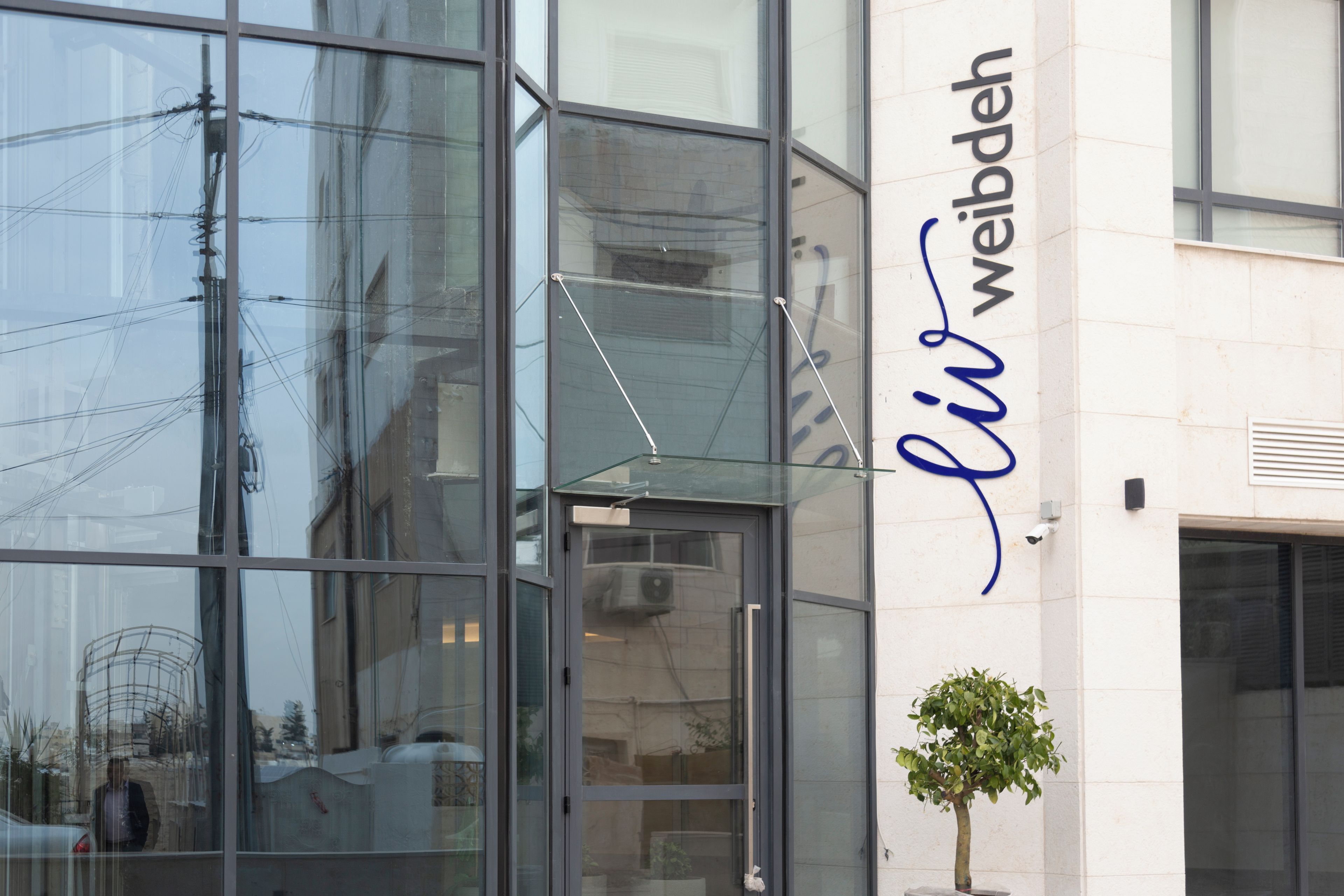 A building with large glass windows reflecting the surroundings, a clear sky, and a sign with the name "li weibdeh".