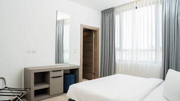 A modern hotel room with a bed, a desk with a mirror, a wardrobe, and a window with curtains.