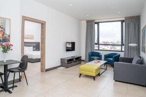 Une chambre d’hôtel moderne équipée d’un lit, d’une télévision, d’un coin repas avec une table et des chaises, d’un canapé et d’une grande fenêtre donnant sur la ville.