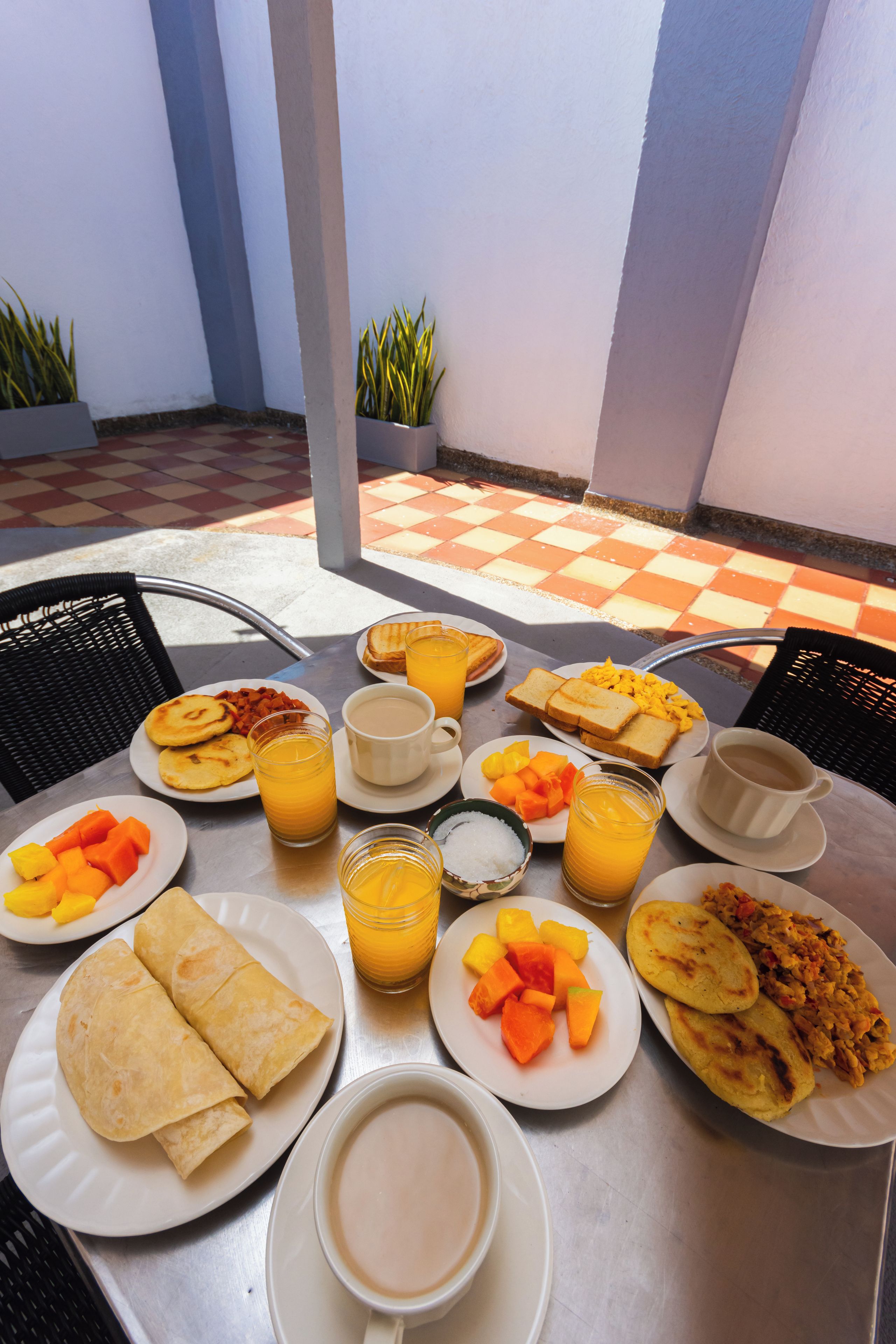 Una tavola imbandita con prodotti per la colazione, tra cui pancake, uova e frutta, con un pavimento a scacchi e piante in vaso sullo sfondo.