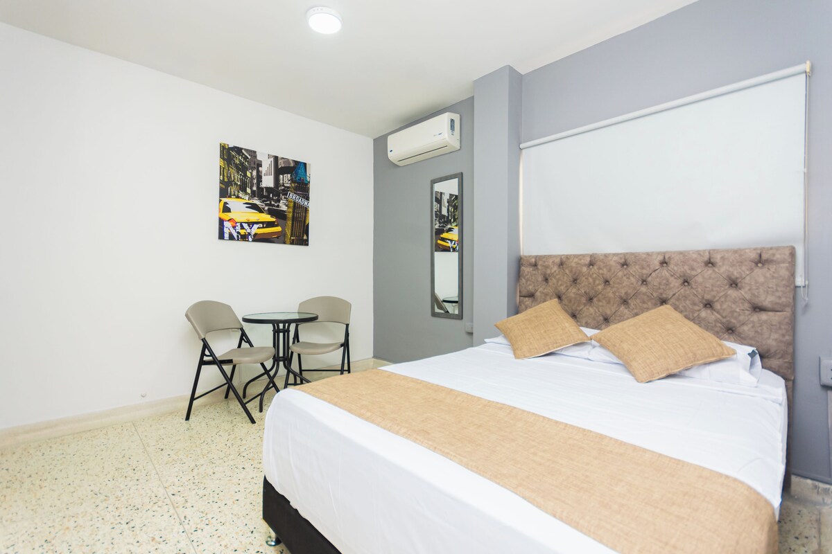 A hotel room with a large bed, a small table, two chairs, and a wall-mounted air conditioner.