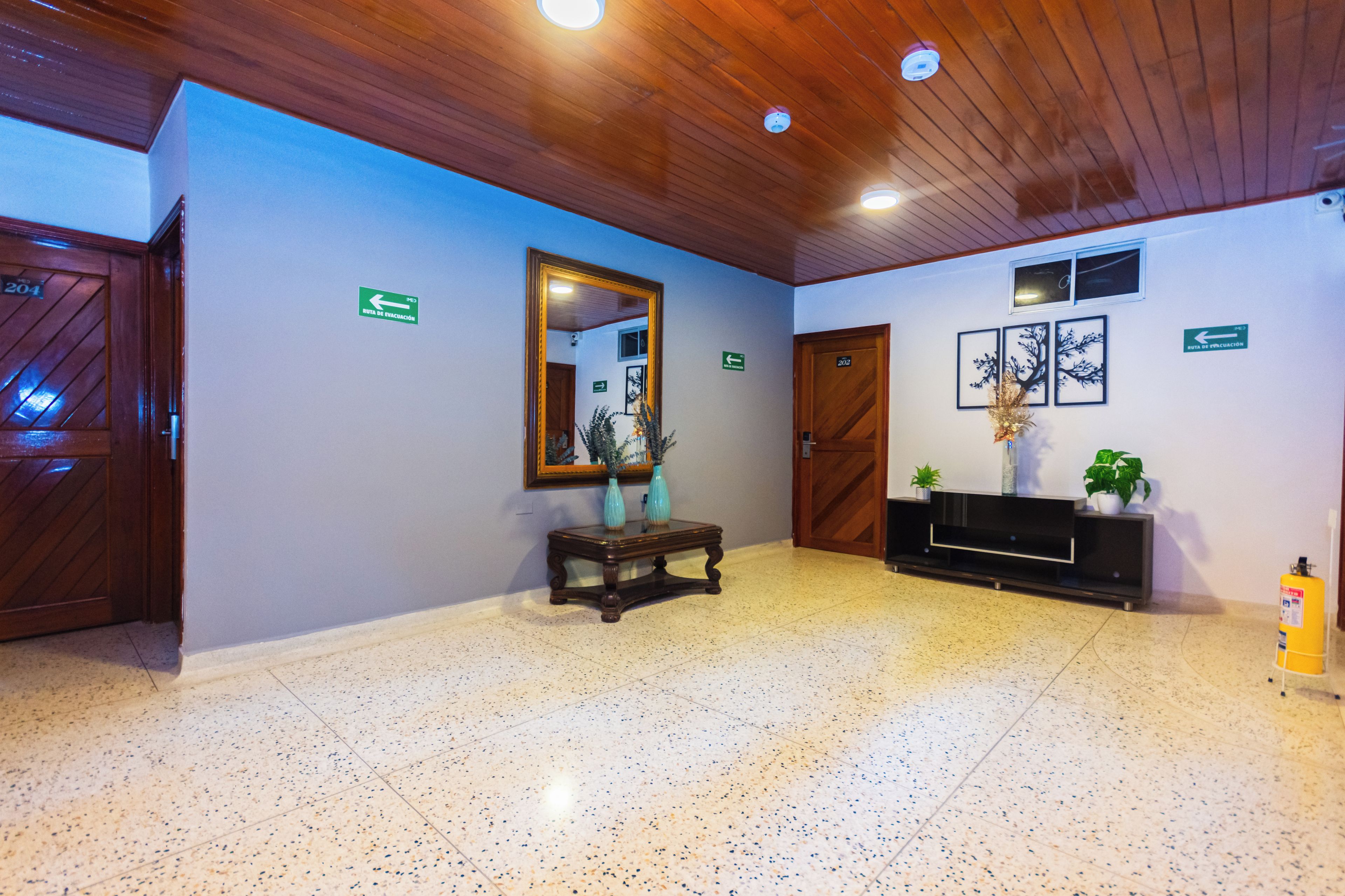 A lobby with a wooden ceiling, a mirror, a console table with vases, a fire extinguisher, and directional signs.