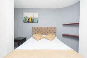 A modern bedroom with a tufted headboard, a bed with two pillows, a small table, and a cityscape painting on the wall. - Suite 94 Hotel Baq (Barranquilla)