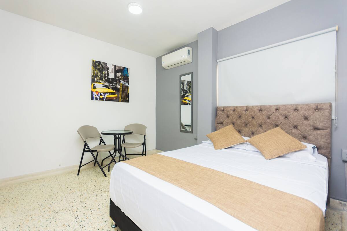 Een hotelkamer met een groot bed, een kleintje tafeltje, twee stoelen en een airconditioner aan de muur.