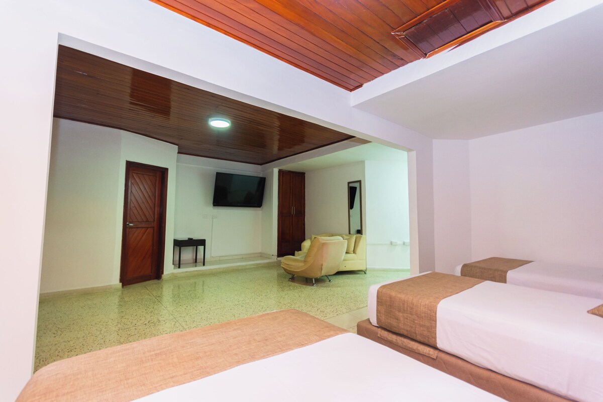 A hotel room with a wooden ceiling, a TV mounted on the wall, a sofa, two beds, and a small table.