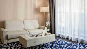A hotel room with a sofa, a coffee table with a magazine, a lamp, and a window with sheer curtains. - Grandhotel Nabokov Spa & Wellness (Marianske Lazne)