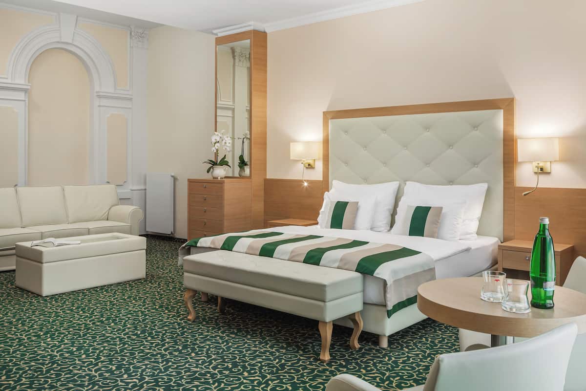 A hotel room with a large bed, a sitting area with a sofa and armchair, a small round table with a bottle and glasses, and a mirror above the bed.