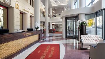 Vestíbulo de un Grand Hotel con mostrador de recepción, alfombra roja con el nombre del hotel, sofá blanco y un ventanal con vistas a la calle.