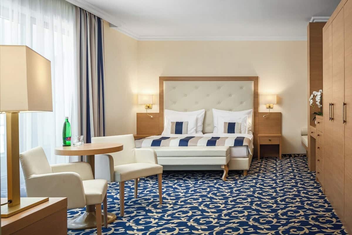 A hotel room with a bed, two bedside tables, a desk with a chair, a wardrobe, and a patterned carpet.