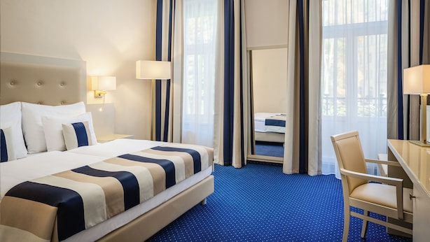 A hotel room with a large bed, a desk, a chair, and a view of the outdoors through the window. - Grandhotel Nabokov Spa & Wellness (Marianske Lazne)